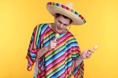 Photo of Young man in Mexican sombrero hat and poncho with maracas on yellow background