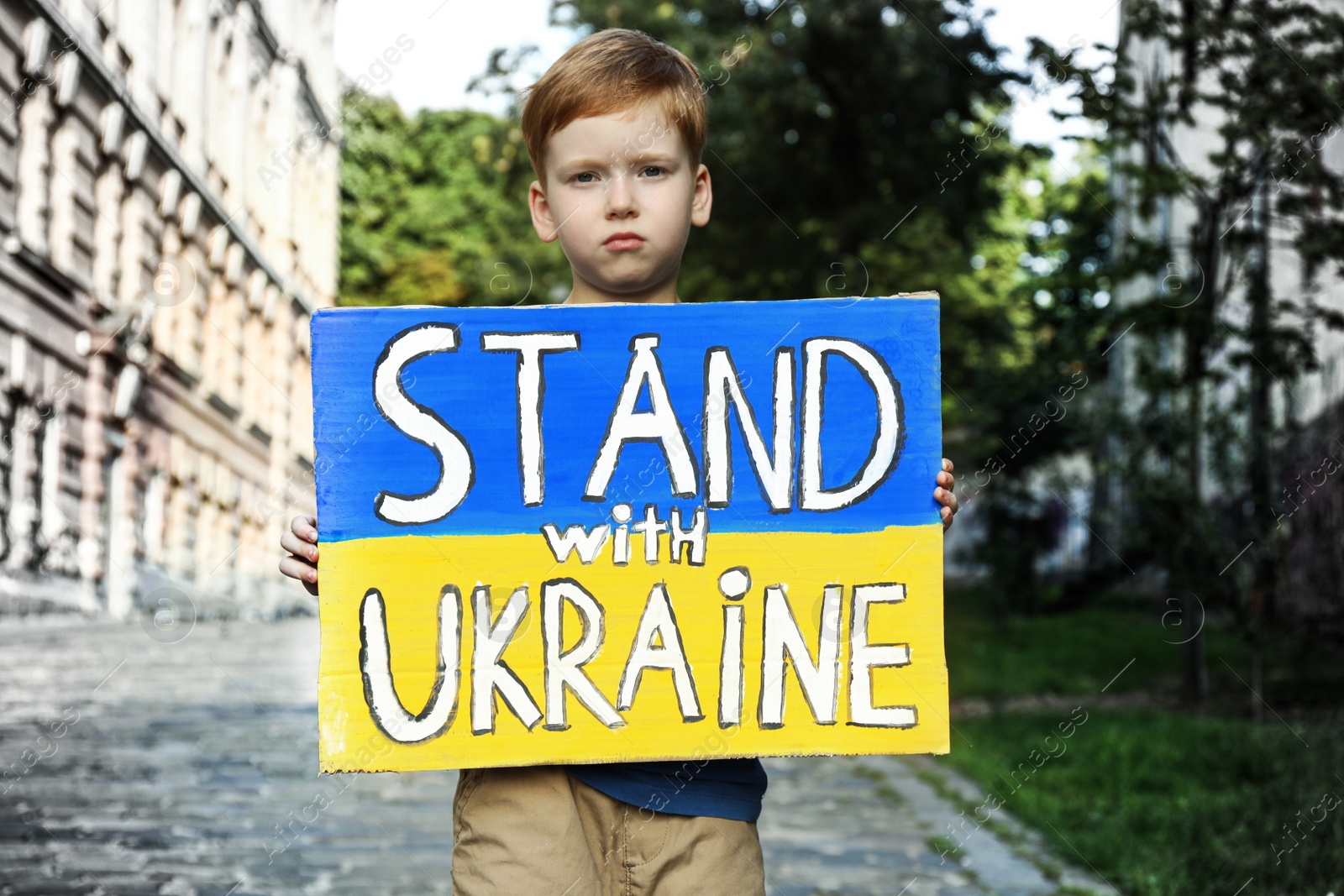Photo of Sad boy holding poster in colors of national flag and words Stand with Ukraine on city street