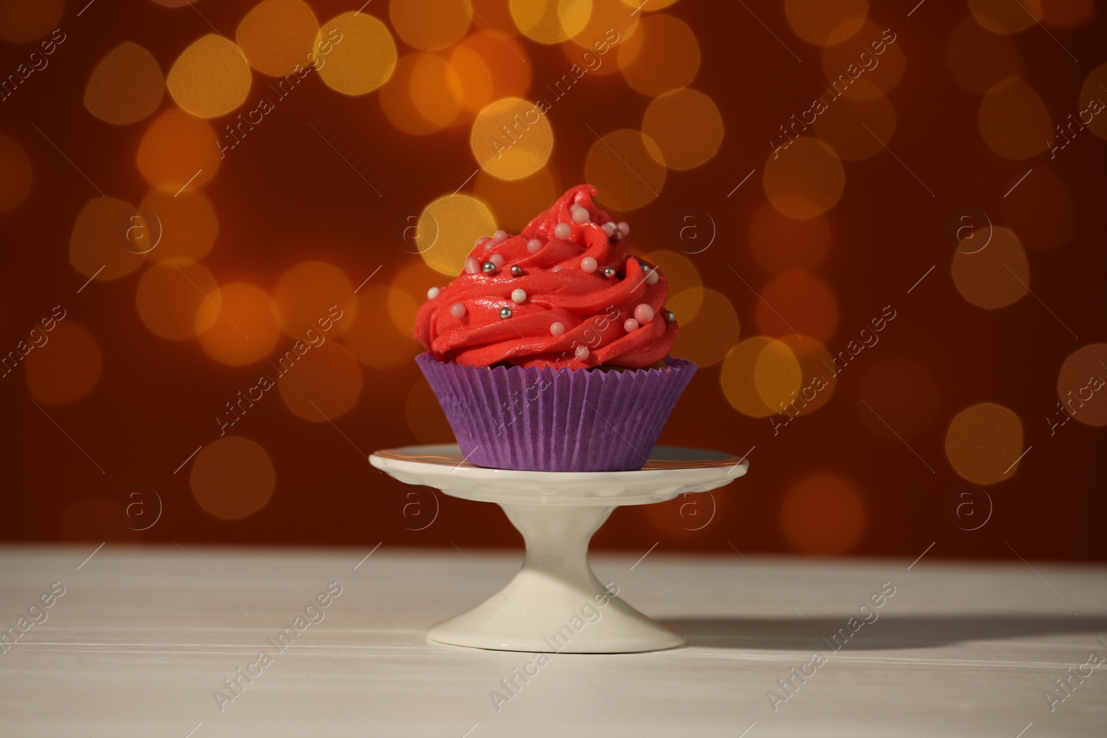 Photo of Stand with tasty cupcake on white table against blurred lights