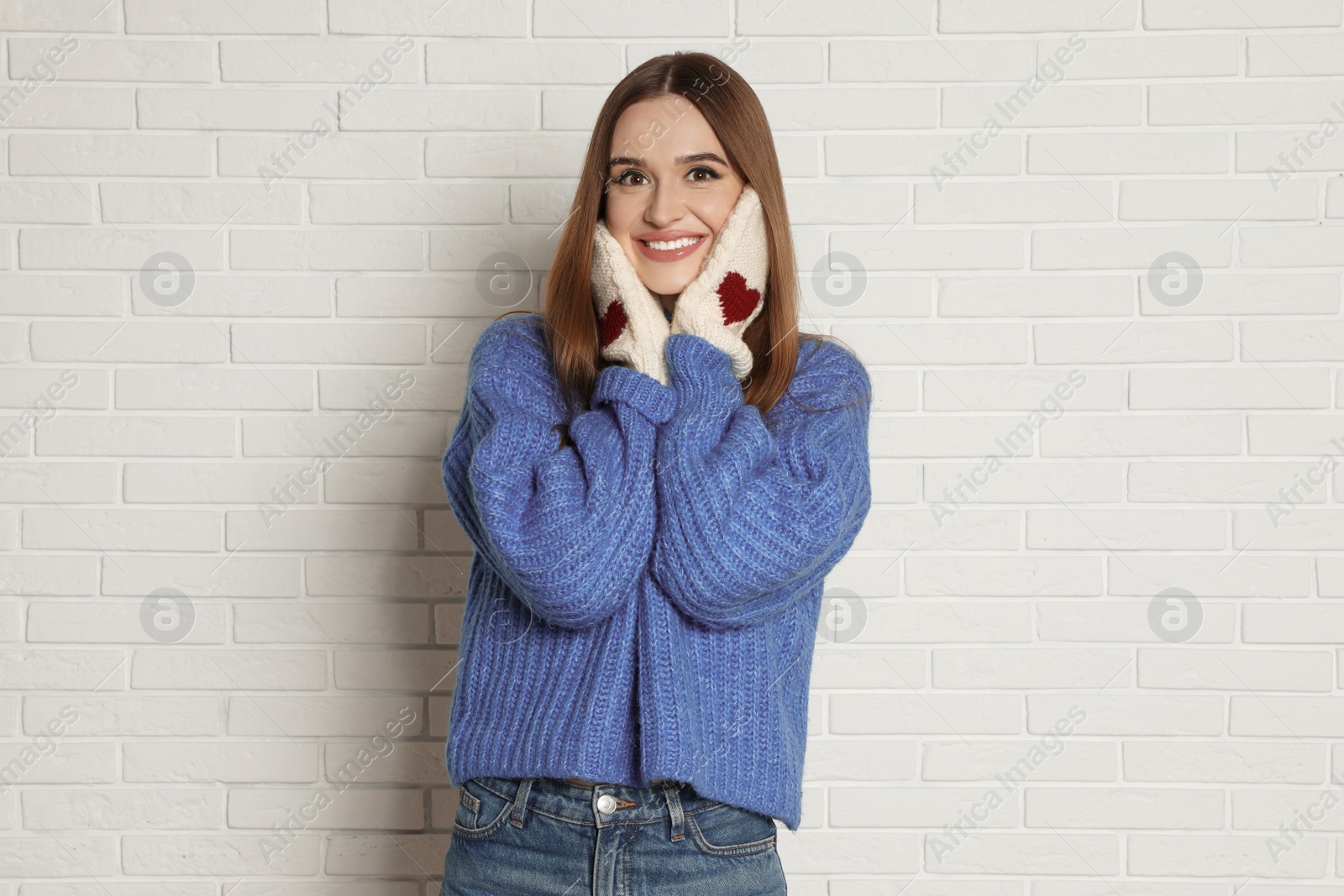 Photo of Beautiful young woman in mittens and blue sweater near white brick wall. Winter season