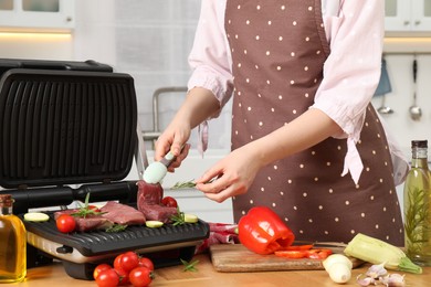 Woman cooking different products with electric grill at wooden table in kitchen, closeup