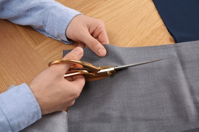 Photo of Man cutting grey fabric with scissors at wooden table, closeup
