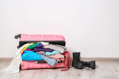 Photo of Suitcase with clothes and boots on floor against white wall, space for text. Winter vacation