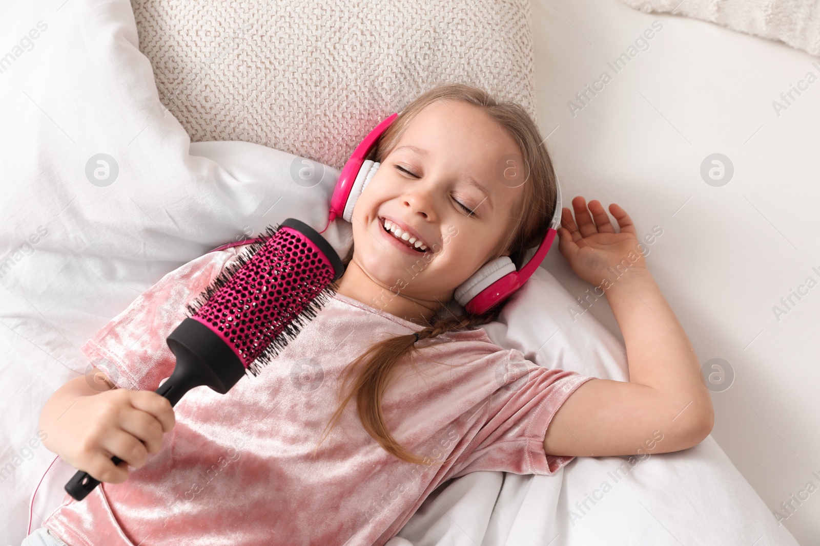 Photo of Cute little girl in headphones with hairbrush singing on bed