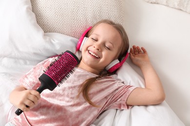 Photo of Cute little girl in headphones with hairbrush singing on bed