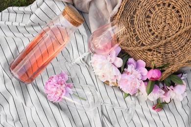 Photo of Bottle and glasses of rose wine near straw bag with beautiful peonies on light blanket, flat lay