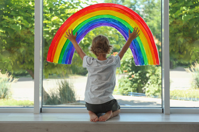 Little boy near rainbow painting on window indoors, back view. Stay at home concept