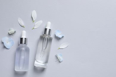 Bottles of cosmetic serums and beautiful flowers on light grey background, flat lay. Space for text