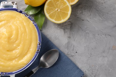 Delicious lemon curd in bowl, fresh citrus fruit, green leaves and spoon on grey table, top view. Space for text