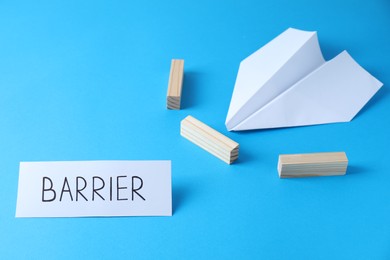 Photo of Paper plane movement blocked by wooden blocks and card with word Barrier on light blue background. Development through obstacles overcoming