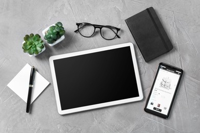 Photo of Online store website on device screen. Tablet, smartphone, stationery, glasses and houseplants on grey table, flat lay