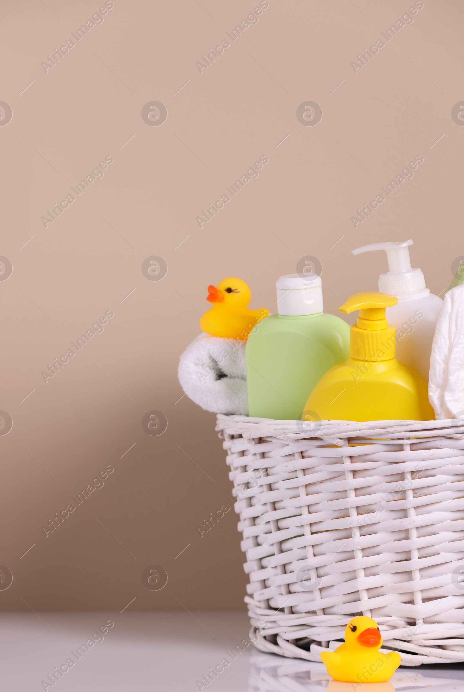 Photo of Wicker basket with baby cosmetic products, bath accessories and rubber ducks on white table against beige background