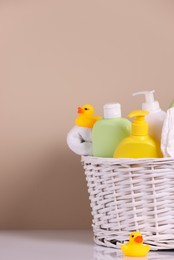 Photo of Wicker basket with baby cosmetic products, bath accessories and rubber ducks on white table against beige background