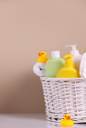 Wicker basket with baby cosmetic products, bath accessories and rubber ducks on white table against beige background