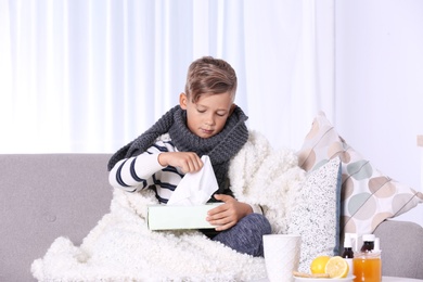 Photo of Ill little boy suffering from cough with box of tissues on sofa