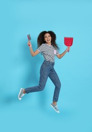 Photo of African American woman with broom and dustpan jumping on light blue background