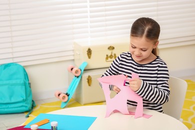 Cute little girl cutting pink paper at desk in room. Home workplace