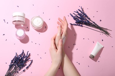 Woman applying hand cream and lavender flowers on pink background, top view