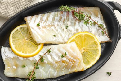 Photo of Fresh raw cod fillets with thyme and lemon in baking dish on white table, top view