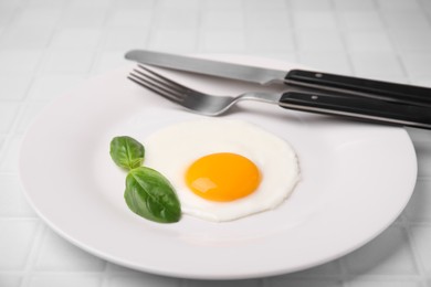 Photo of Tasty fried egg with basil in plate on white tiled table, closeup