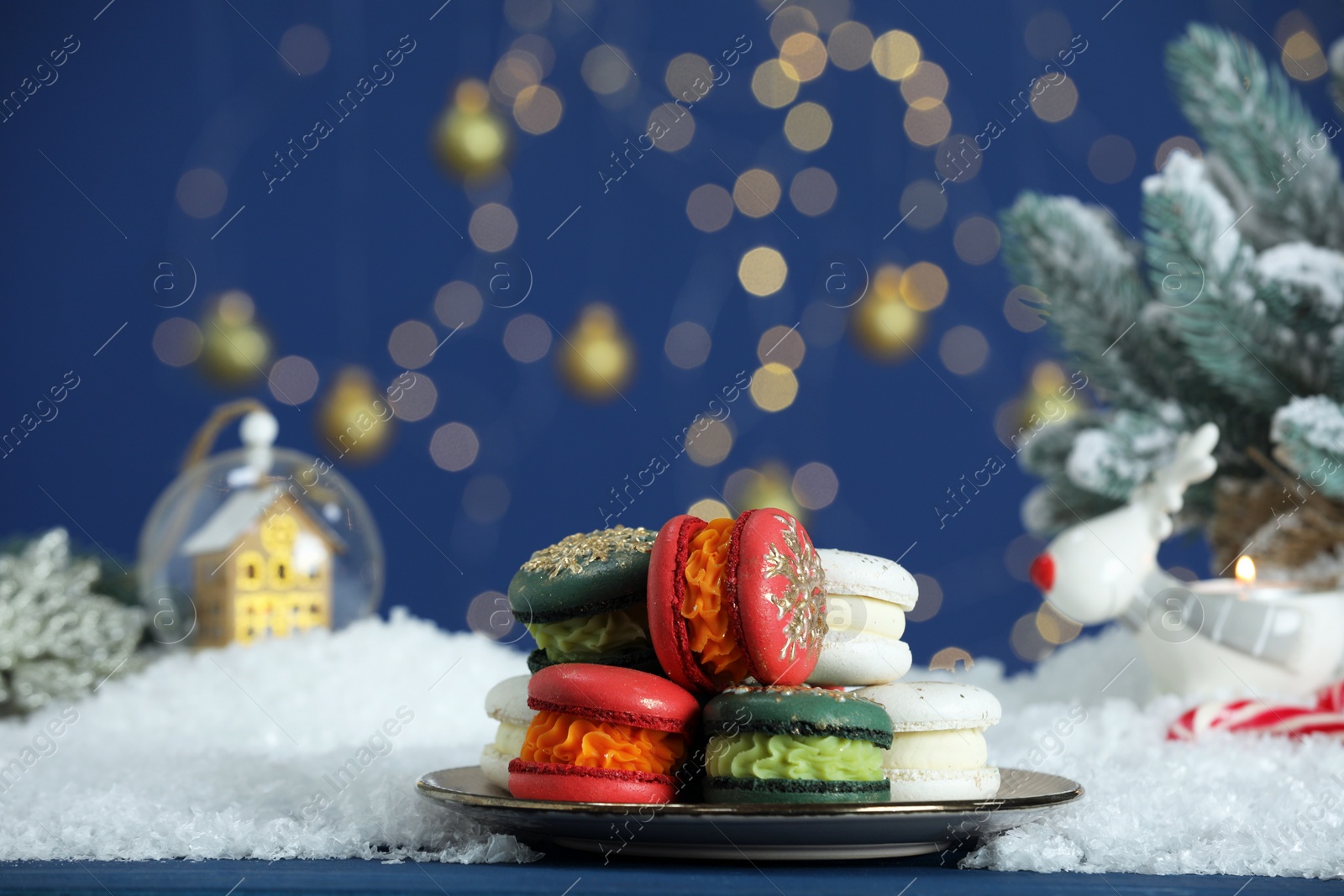 Photo of Different decorated Christmas macarons on blue table with artificial snow against blurred lights