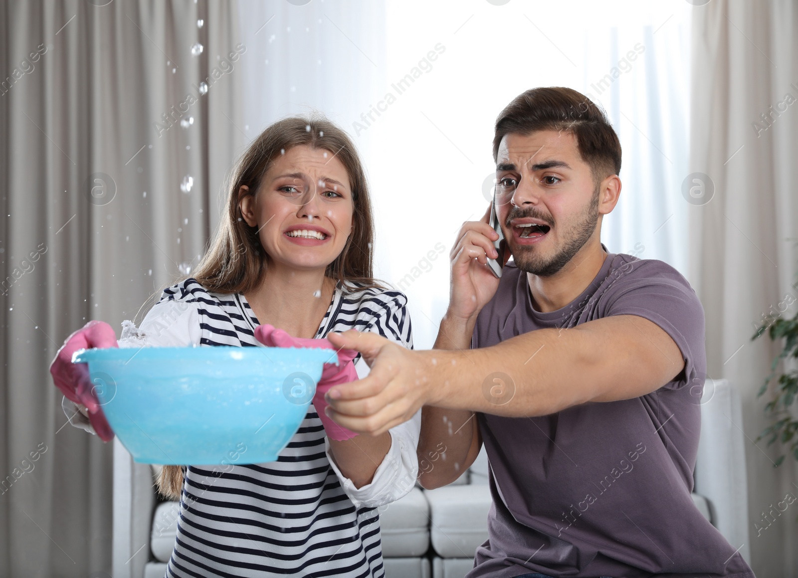 Photo of Emotional young woman collecting water leaking from ceiling while her husband calling plumber in living room