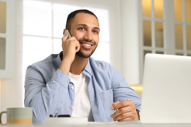 Photo of Young man talking on smartphone while working with laptop at desk in kitchen. Home office