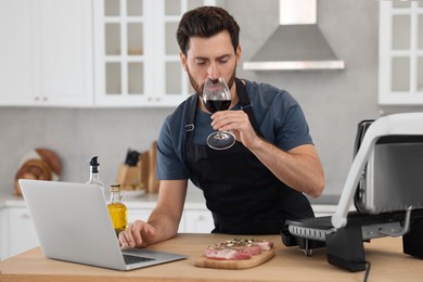 Photo of Man with glass of wine making dinner while watching online cooking course via laptop in kitchen