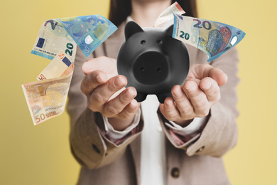 Young woman holding piggy bank and Euro banknotes falling on color background, closeup 