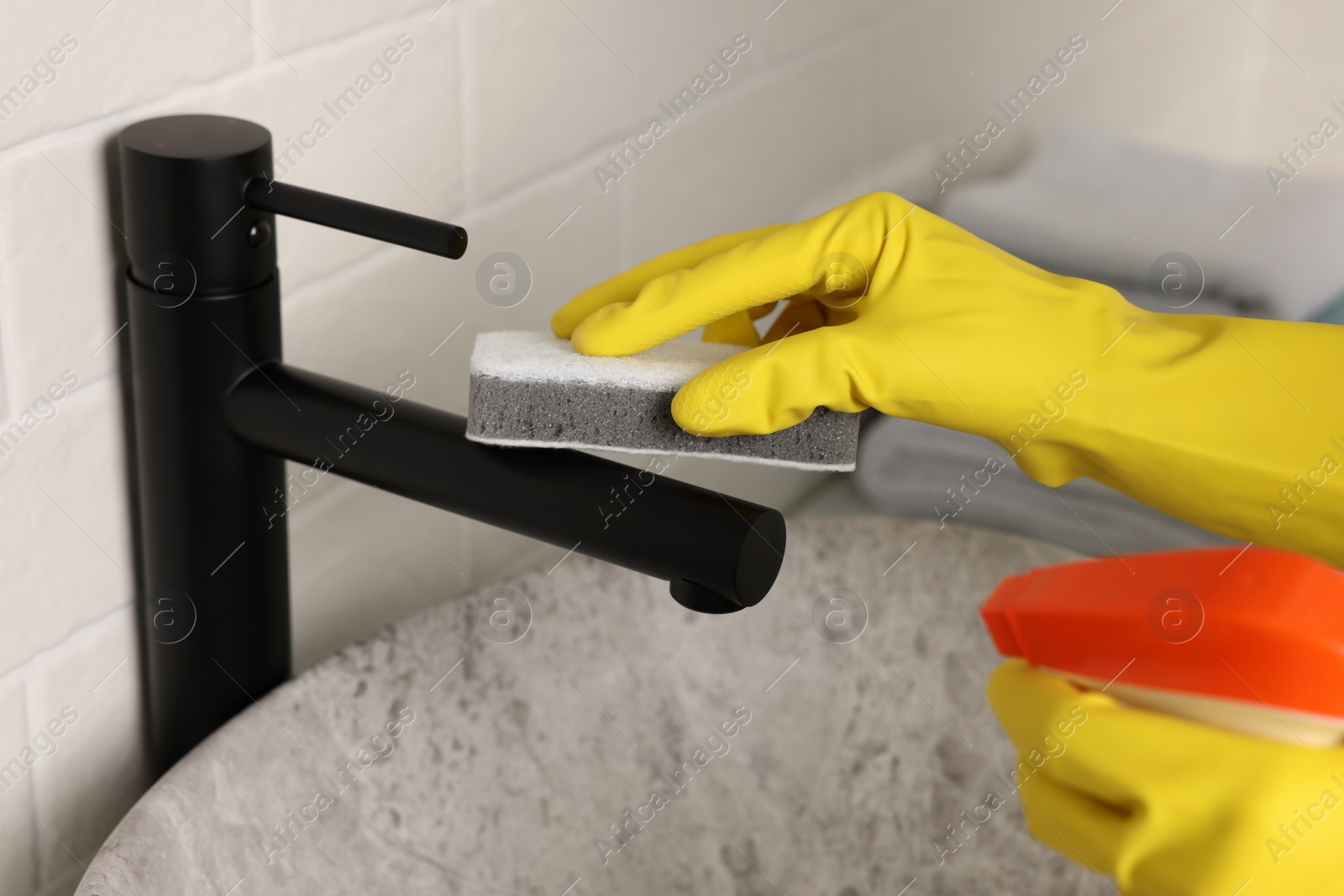 Photo of Woman in gloves cleaning faucet of bathroom sink with sponge, closeup