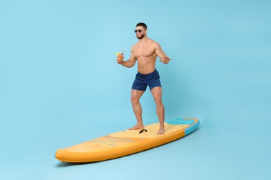 Photo of Happy man with refreshing drink balancing on SUP board against light blue background