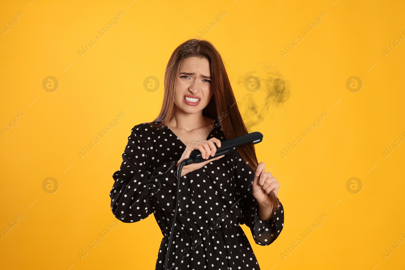 Image of Stressed young woman with flattening iron on yellow background. Hair damage