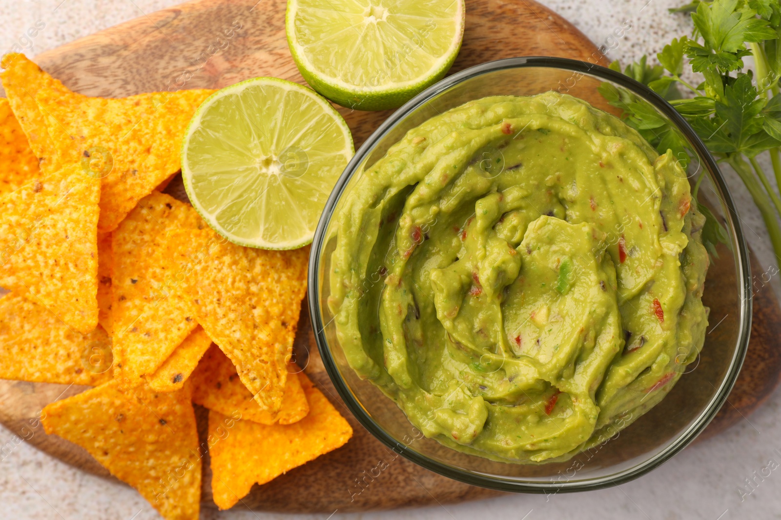 Photo of Wooden board with bowl of delicious guacamole, nachos chips and lime on white tiled table, top view