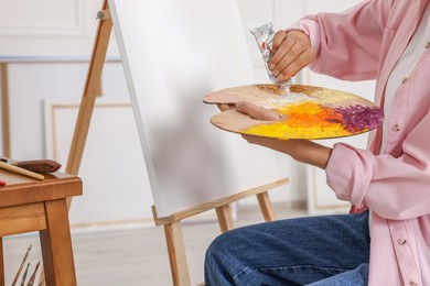 Photo of Woman mixing paints on palette near easel in studio, closeup