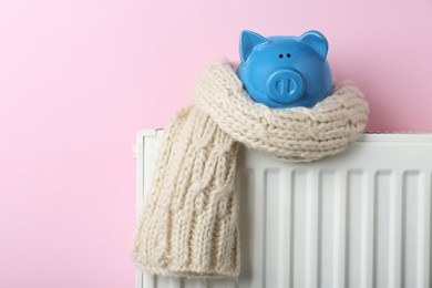 Photo of Piggy bank wrapped in scarf on heating radiator against pink background, space for text