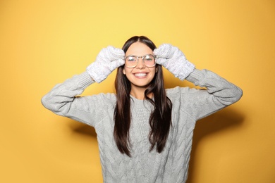 Happy young woman wearing warm sweater and mittens on yellow background 