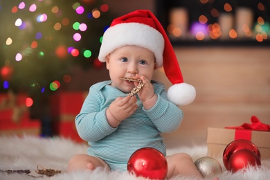 Little baby in Santa hat playing with Christmas decoration against blurred festive lights indoors