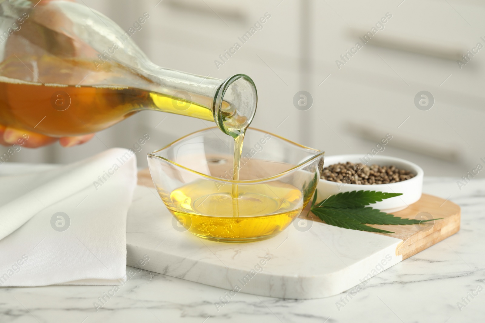 Photo of Woman pouring hemp oil into glass bowl at white marble table, closeup