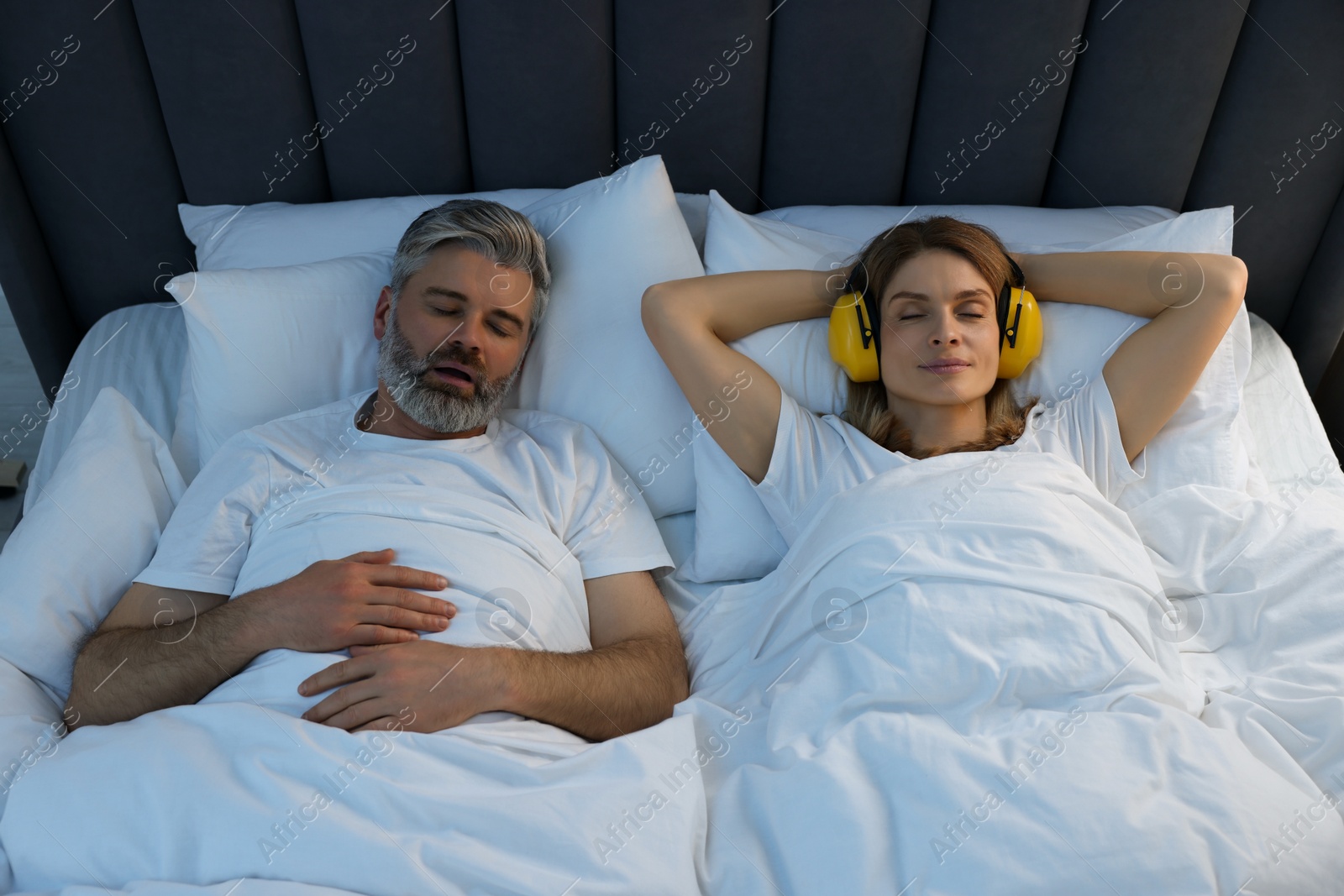 Photo of Smiling woman with headphones lying near her snoring husband in bed at home