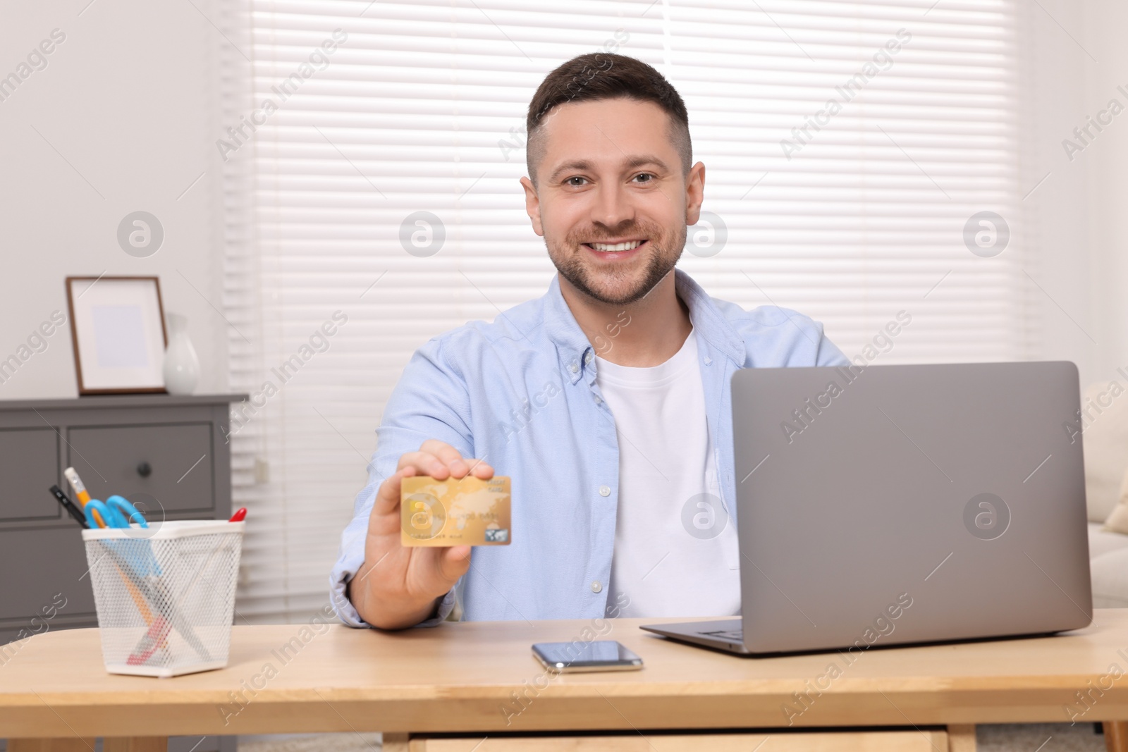 Photo of Handsome man with credit card using laptop for online shopping at wooden table in room