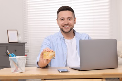 Handsome man with credit card using laptop for online shopping at wooden table in room