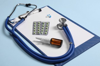 Photo of Stethoscope, pills, nasal spray and clipboard on light blue background, closeup. Medical tool