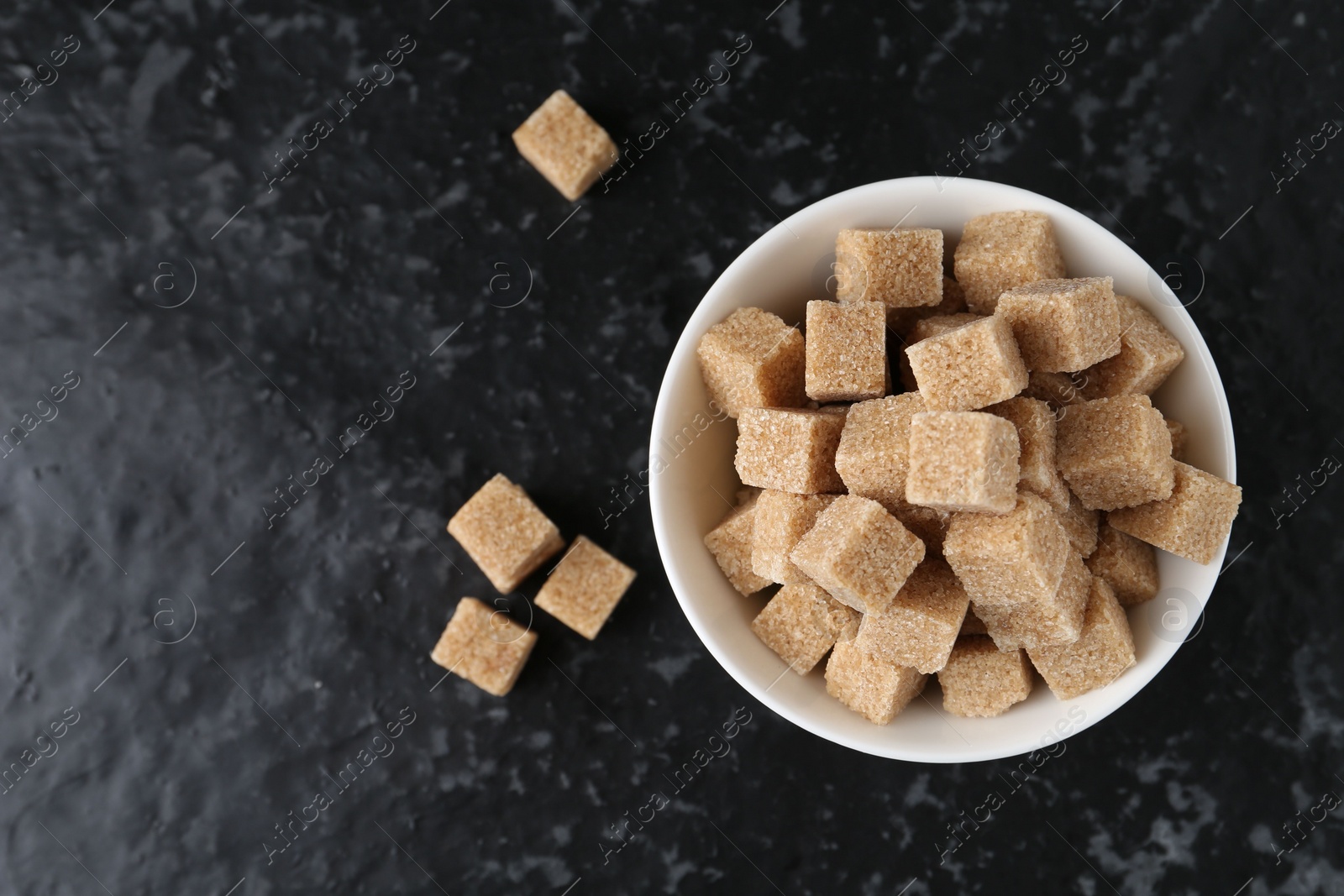 Photo of Brown sugar cubes in bowl on dark textured table, top view. Space for text