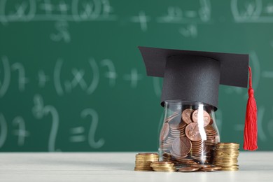 Scholarship concept. Glass jar with coins and graduation cap on white wooden table, space for text