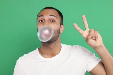 Photo of Portrait of young man blowing bubble gum and showing peace gesture on green background