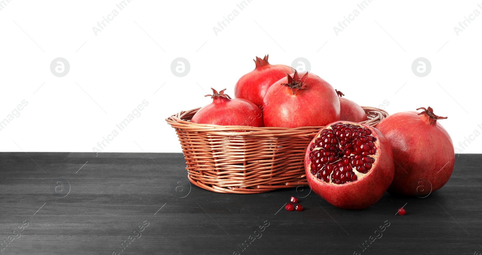 Photo of Fresh pomegranates in wicker basket on black wooden table against white background, space for text