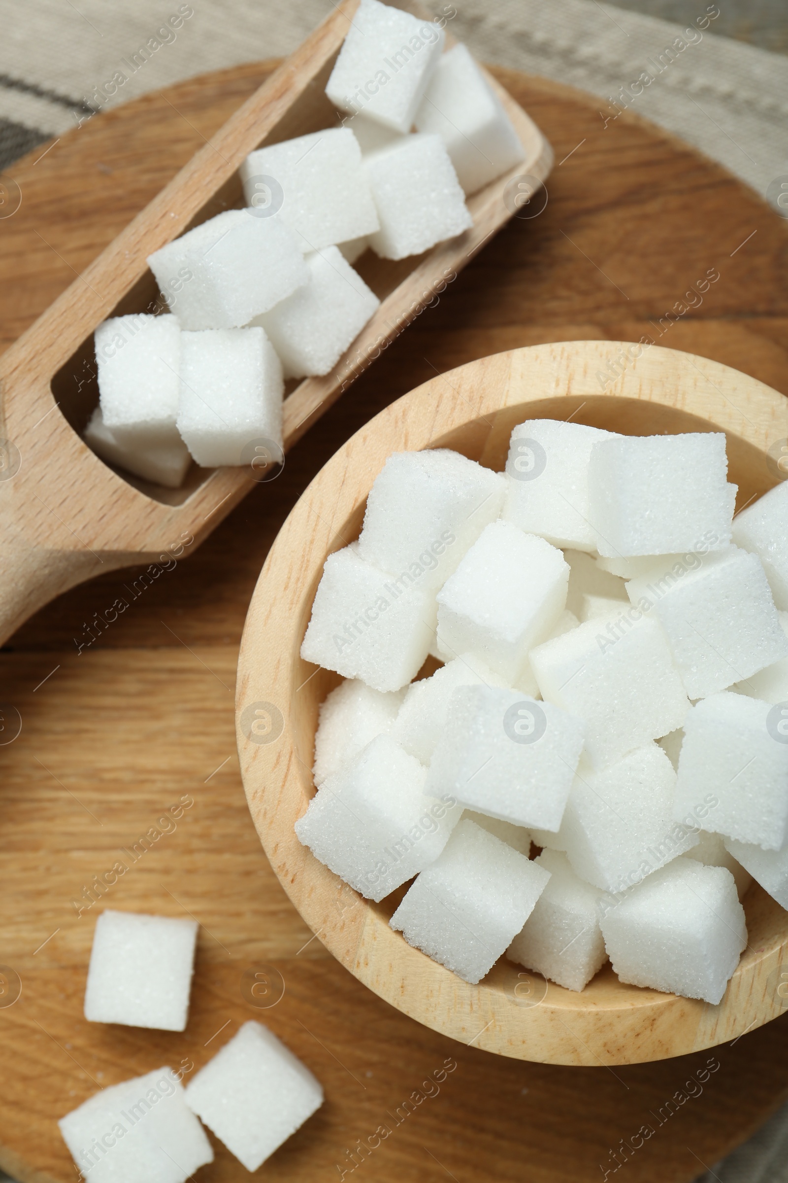 Photo of White sugar cubes in bowl and scoop on wooden board, top view