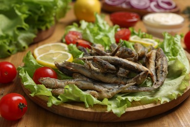 Photo of Delicious fried anchovies served with lemon, tomatoes and lettuce leaves on wooden table, closeup