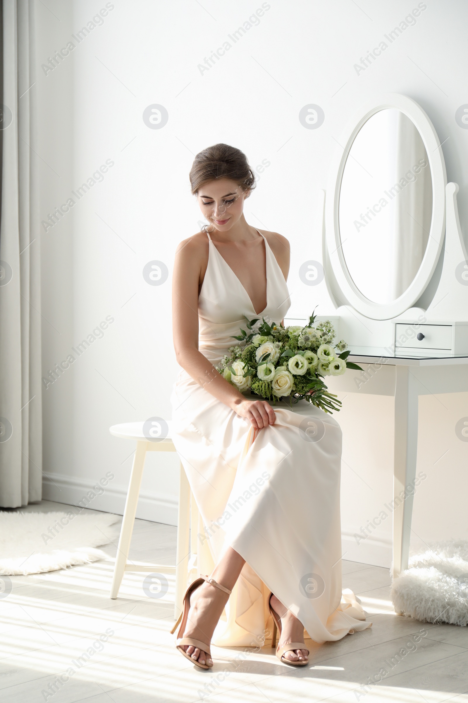 Photo of Young bride in wedding dress with beautiful bouquet near mirror indoors