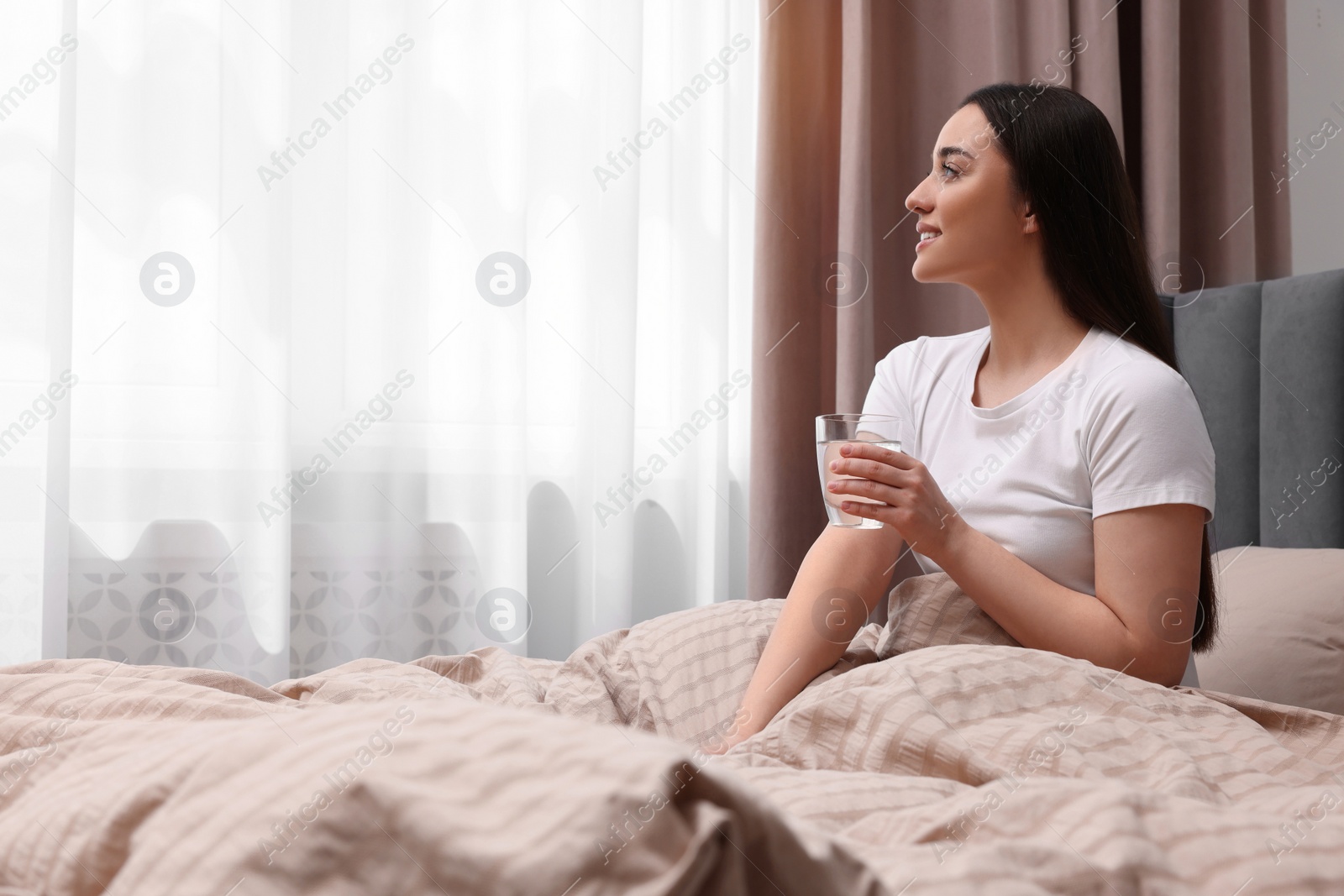 Photo of Beautiful woman with glass of water on bed at home, space for text. Lazy morning